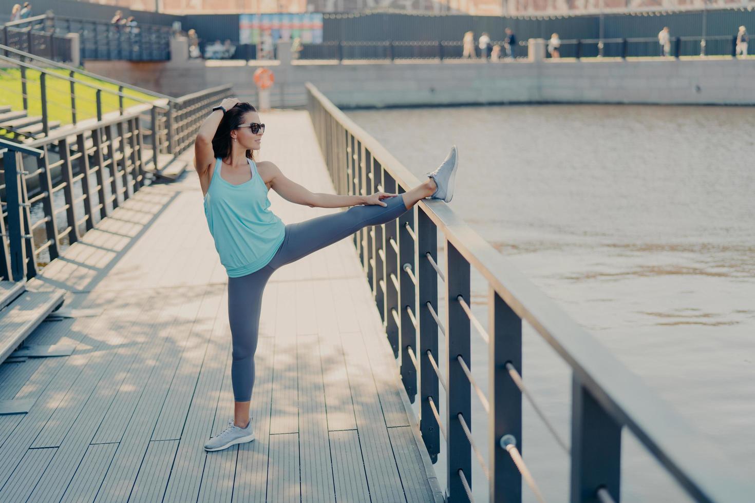Young fit woman with dark hair stretches legs on fence warms up before jogging wears sunglasses t shirt leggings and sneakers prepares for cardio training poses outdoor being in good physical shape photo