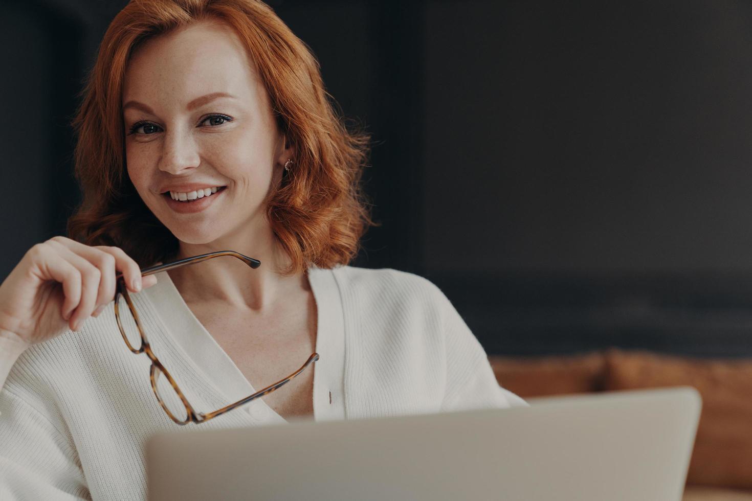 Pleased ginger female student spends time on online education, uses laptop computer and wireless connection, concentrated on remote job, holds spectacles, smiles toothily, installs new application photo