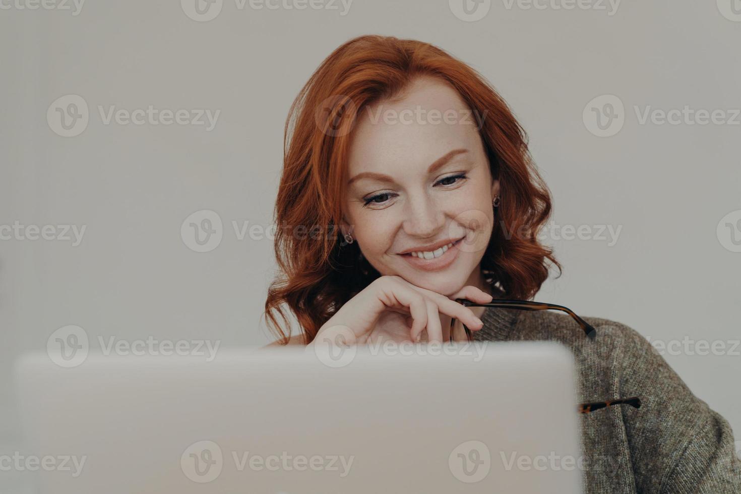 Successful female student with ginger hair and pleasant smile watches interesting tutorial video or webinar, uses laptop computer for e learning, reads information online, keeps hand under chin photo