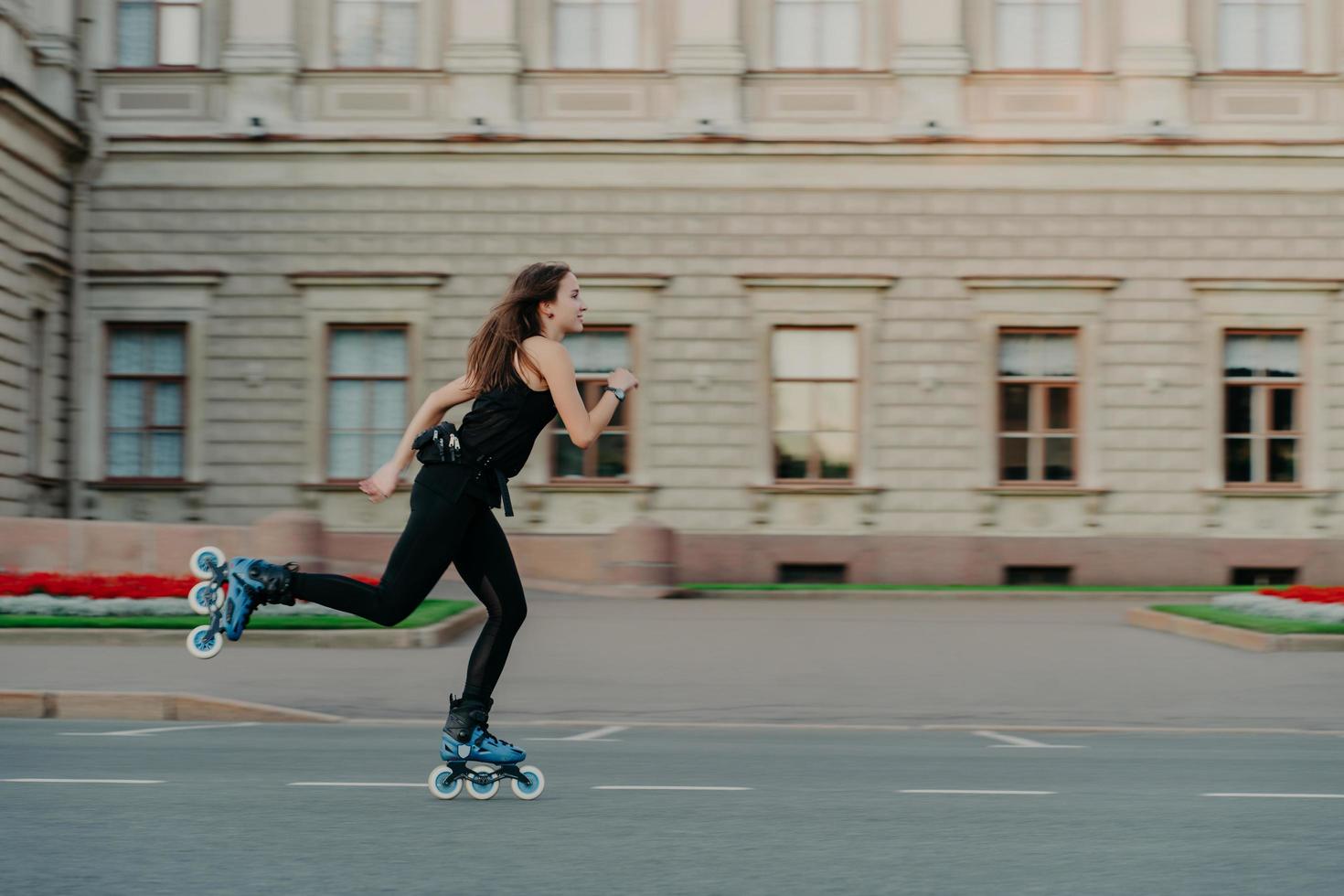 Outdoor physical activities. Healthy young woman has slim fit figure leads active lifestyle spends free time outside rides on rollerblades poses alone in open air against building background photo