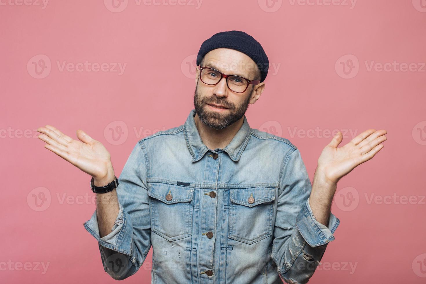 Portrait of hesitant bearded male with doubtful expression, shrugs shoulders, wears denim jacket, glasses and hat, isolated over pink studio background. Confused middle aged man poses indoor photo