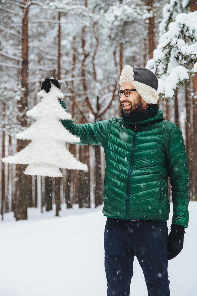 Vertical portrait of dlightful positive bearded male dressed in warm winter clothes, looks with happy expression at artificial fir tree, demonstrates that he protects nature and doesnt cut down trees photo