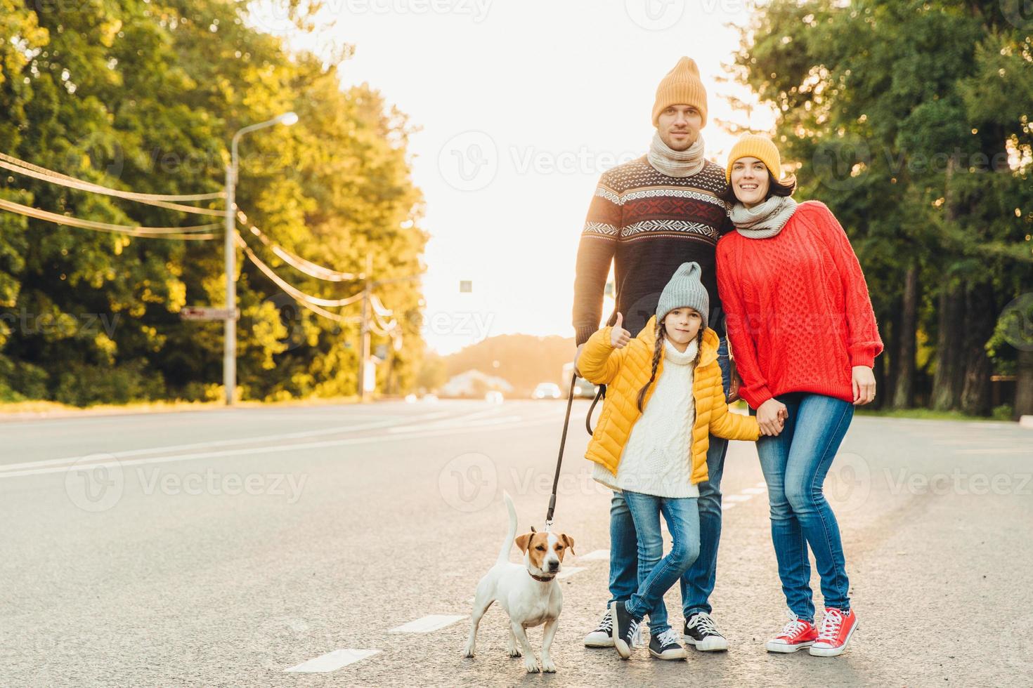 Happy family wear warm clothes walk with dog on road, stand close to each other as pose in camera. Little girl shows ok sign as being glad to spend free time with her parents and favourite pet photo