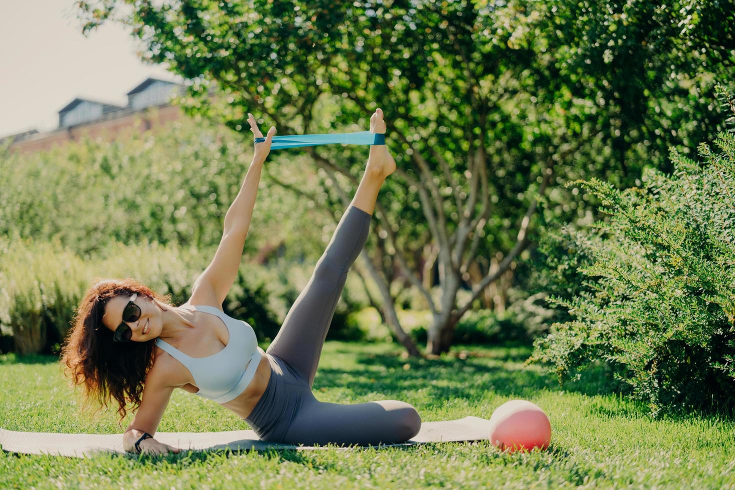 Strong fit woman uses fitness gum to stretch legs and arms stands on side plank at karemat dressed in sportswear poses outdoor during summer day everything is green. Exercises on leg muscles photo