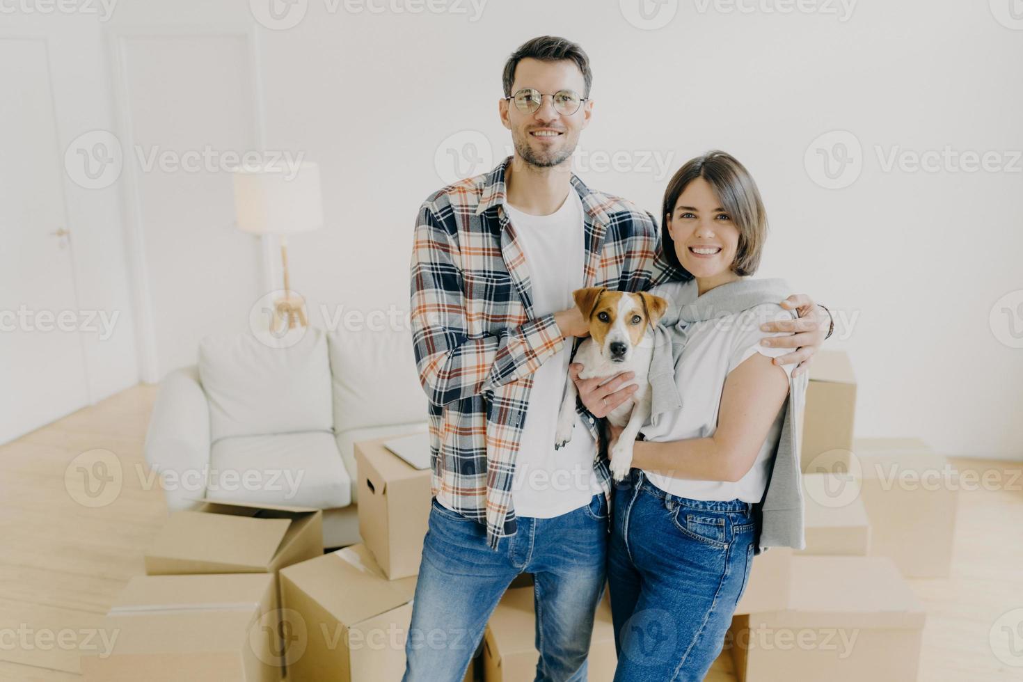 Affectionate family couple celebrate moving day, embrace and holds favourite pet, enjoy relocaton and starting living together, pose in big room among carton boxes. Mortgage and relocation concept photo