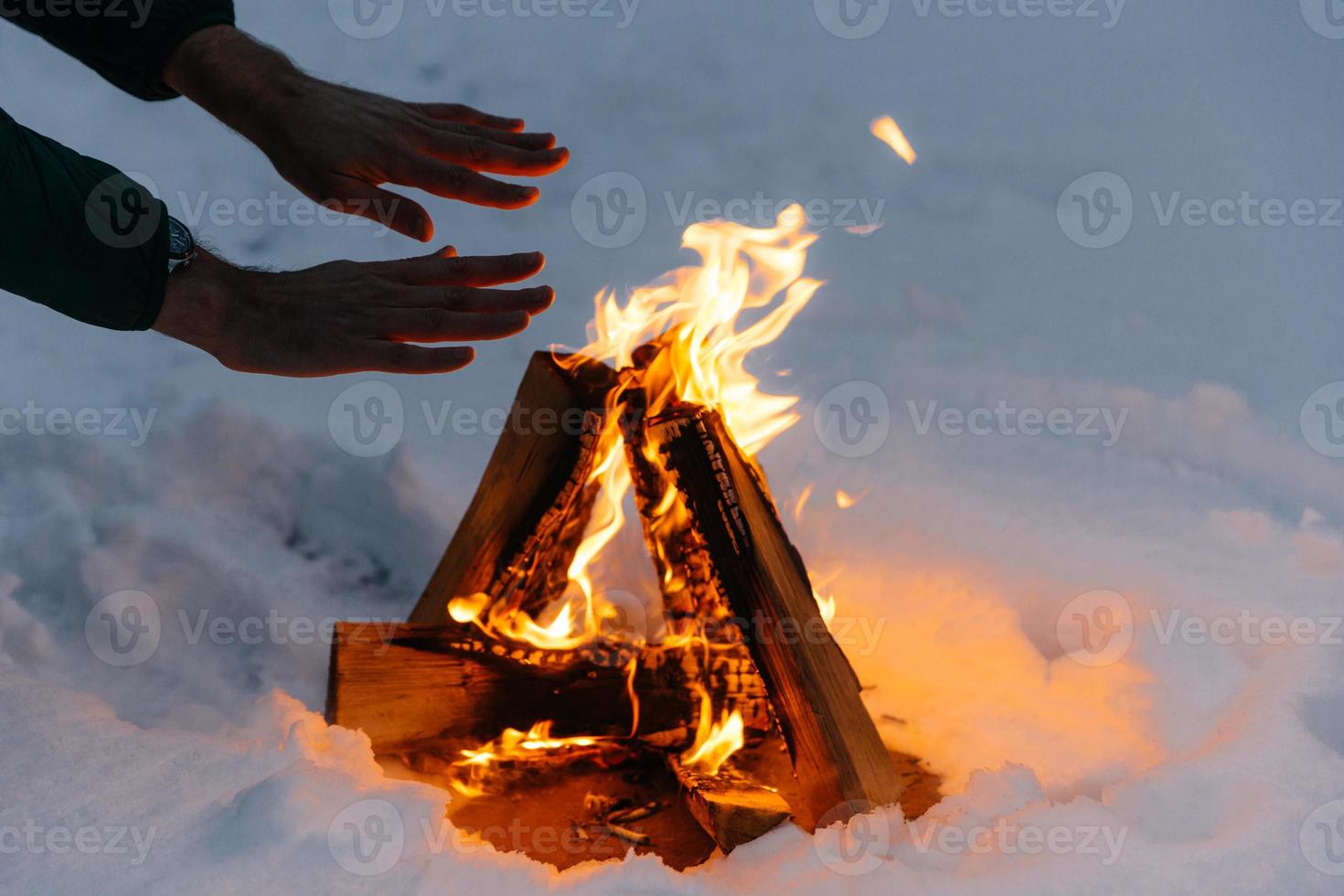Unrecognizable male warms hands on fire in forest during cold winter, tries to warm himself, being cold spend time on cold snow and frosty weather. Burning flame on ground covered with snow. photo