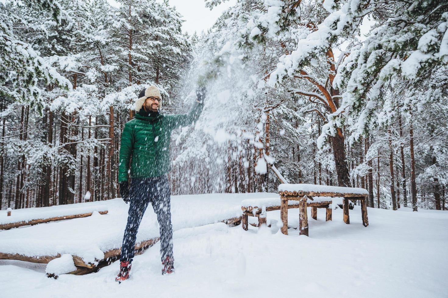 Good looking positive male keeps hand on fir tree, looks with cheerful expression, enjoys beauty of trees covered with snow, spends weekends in forest. Smiling bearded man being in good mood photo