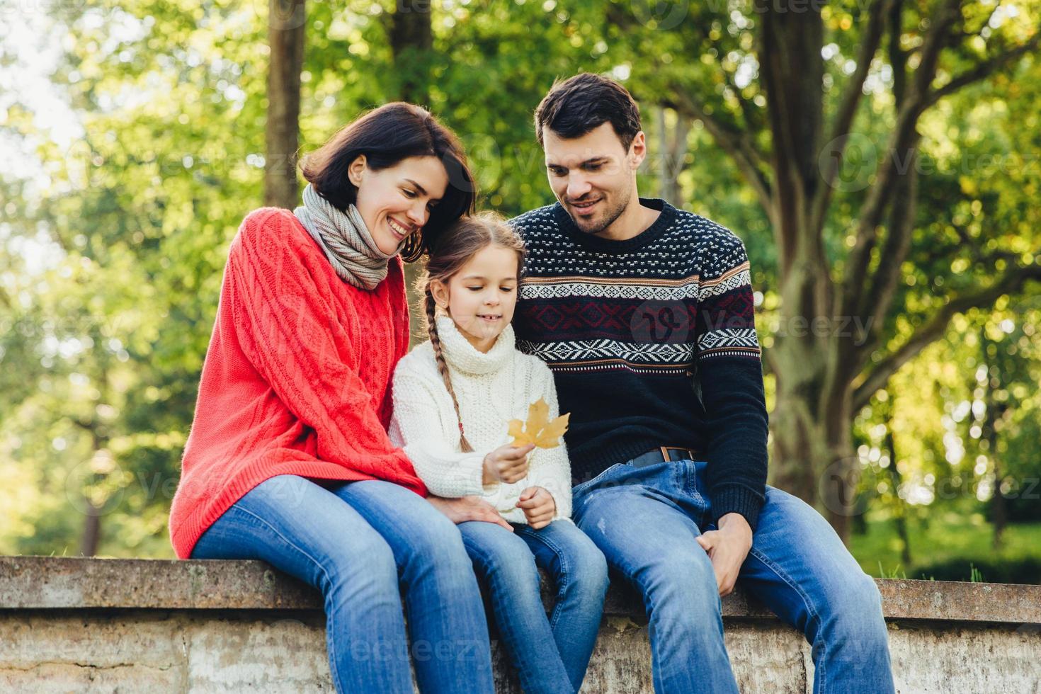 Adorable cute lovely small chld sits between her parents outdoors, holds leaf in hands, spend weekends together. Friendly young family enjoy togetherness, have happy expressions, sit on bridge photo
