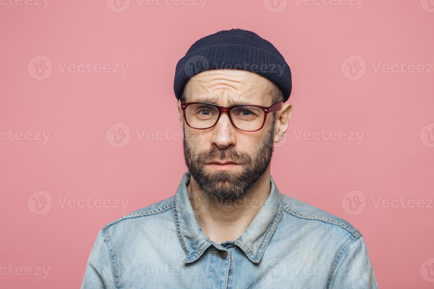 Grumpy bearded man with offended expression, being dissatisfied with something, frowns face, wears spectacles, hat and shirt, isolated over pink background. Displeasure and negativity concept photo