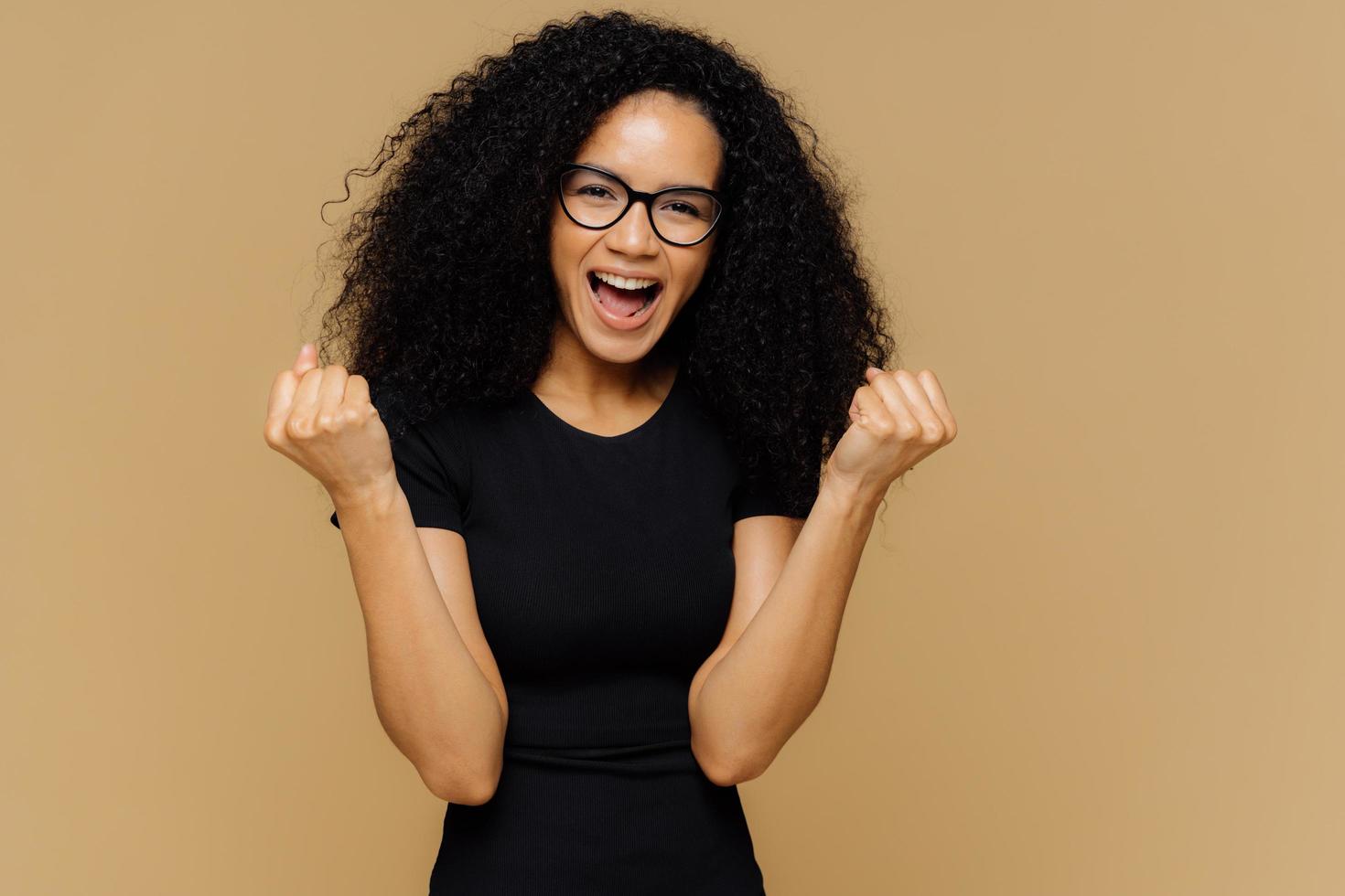 Yeah, I finally did it Optimistic lovely dark skinned curly woman raises clenched fists, exclaims from positive emotions, celebrates victory, dressed in black outfit, isolated over brown background photo