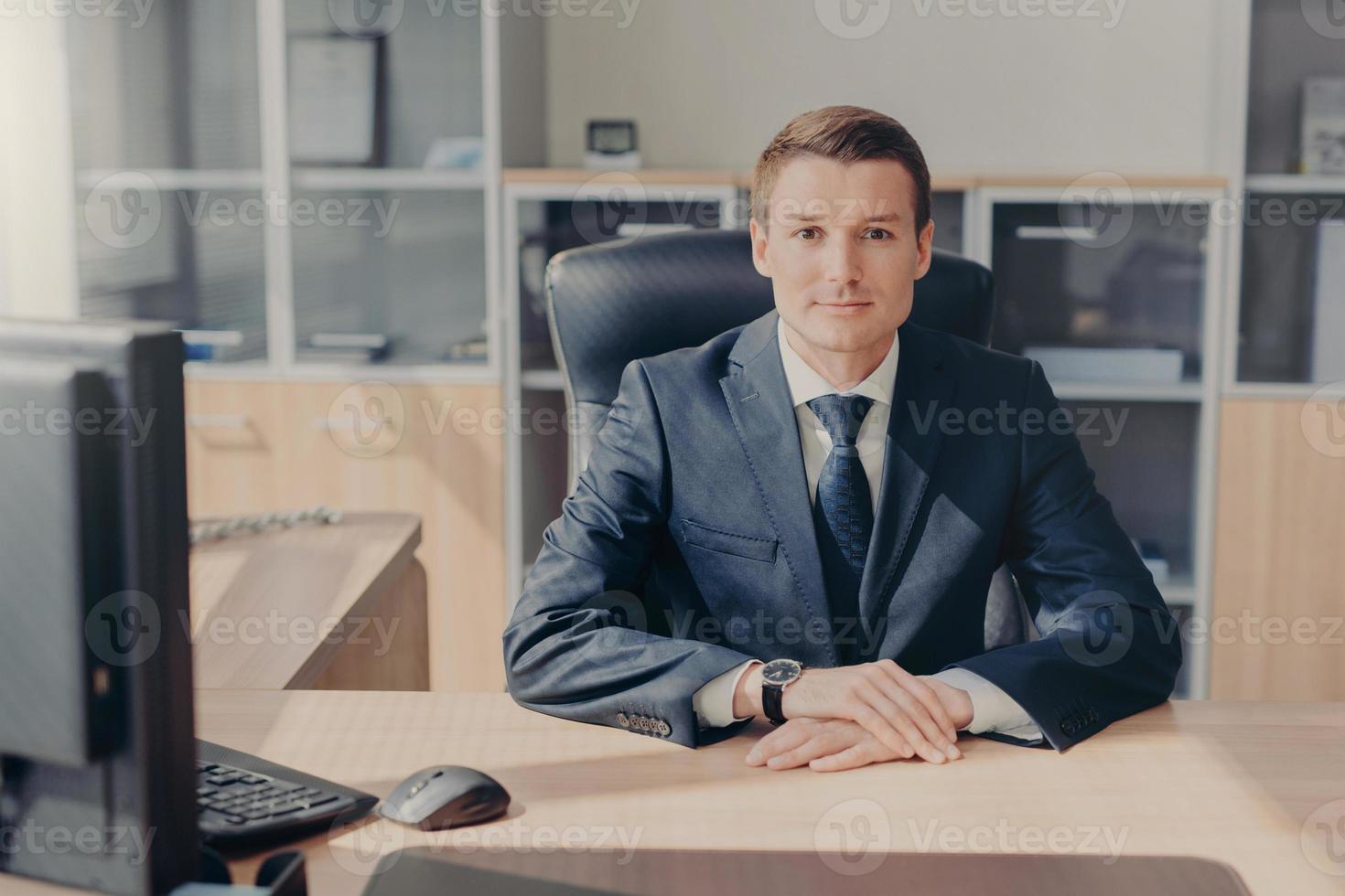 Horizontal shot of prosperous young businessman in elegant suit, sits at work place, surrounded with computer, has confident look directly into camera, being professional in business sphere. photo
