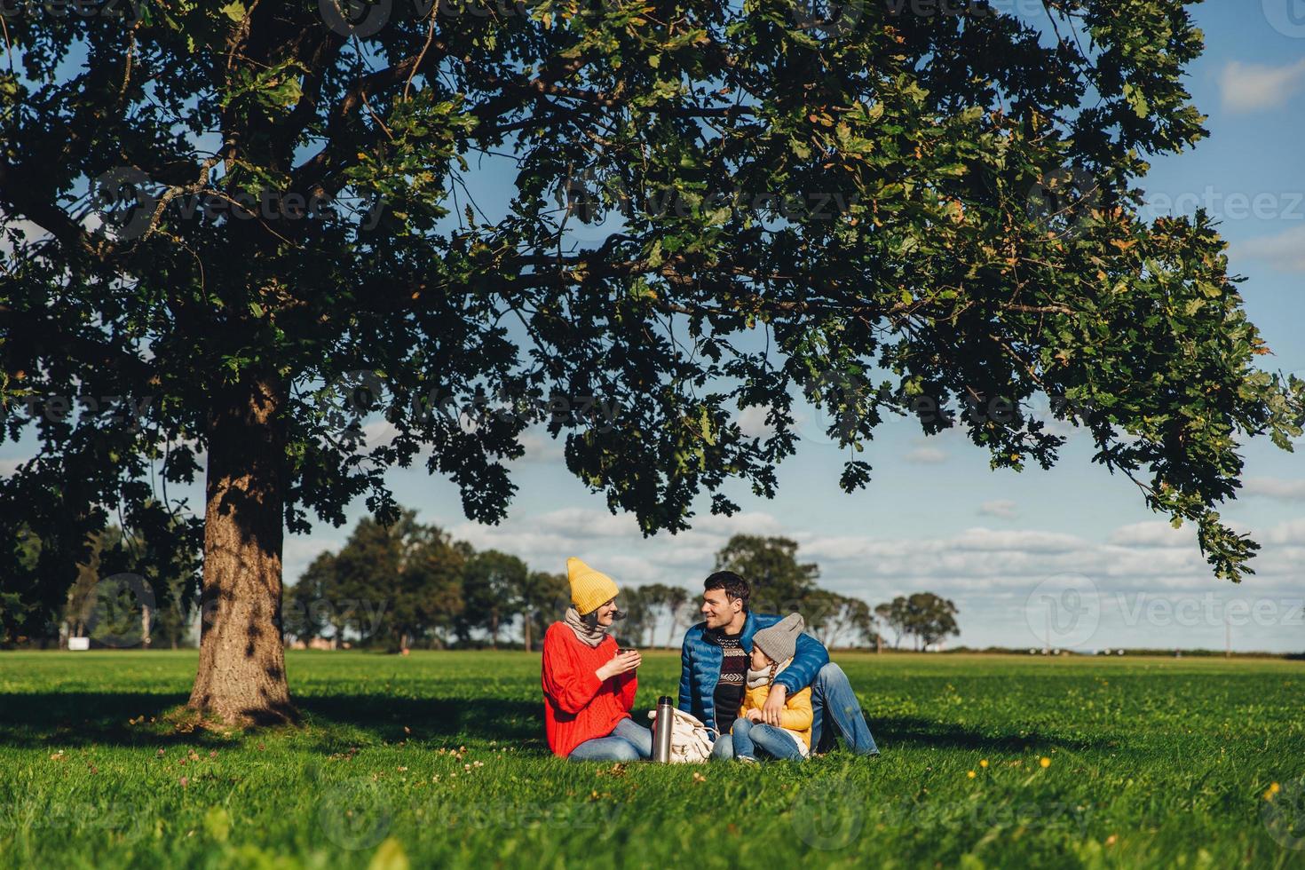Happy family have autumn picnic, sit on green grass, drink hot tea, communicate with each other, enjoy calm atmosphere, admire blue sky and beautiful landscapes. Friendly family members together photo