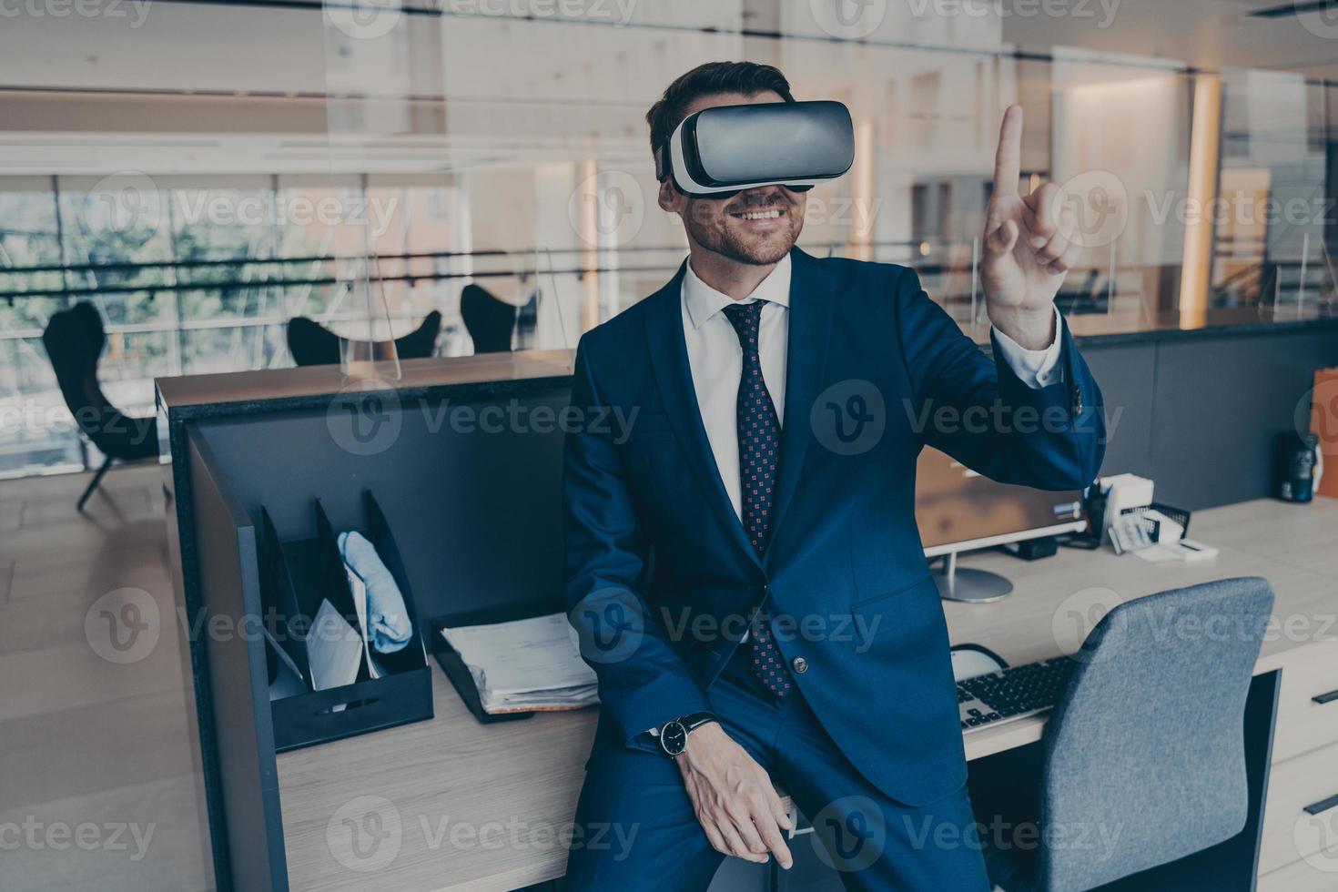Businessman sitting on desk in office and using VR headset on his head, pointing with forefinger photo
