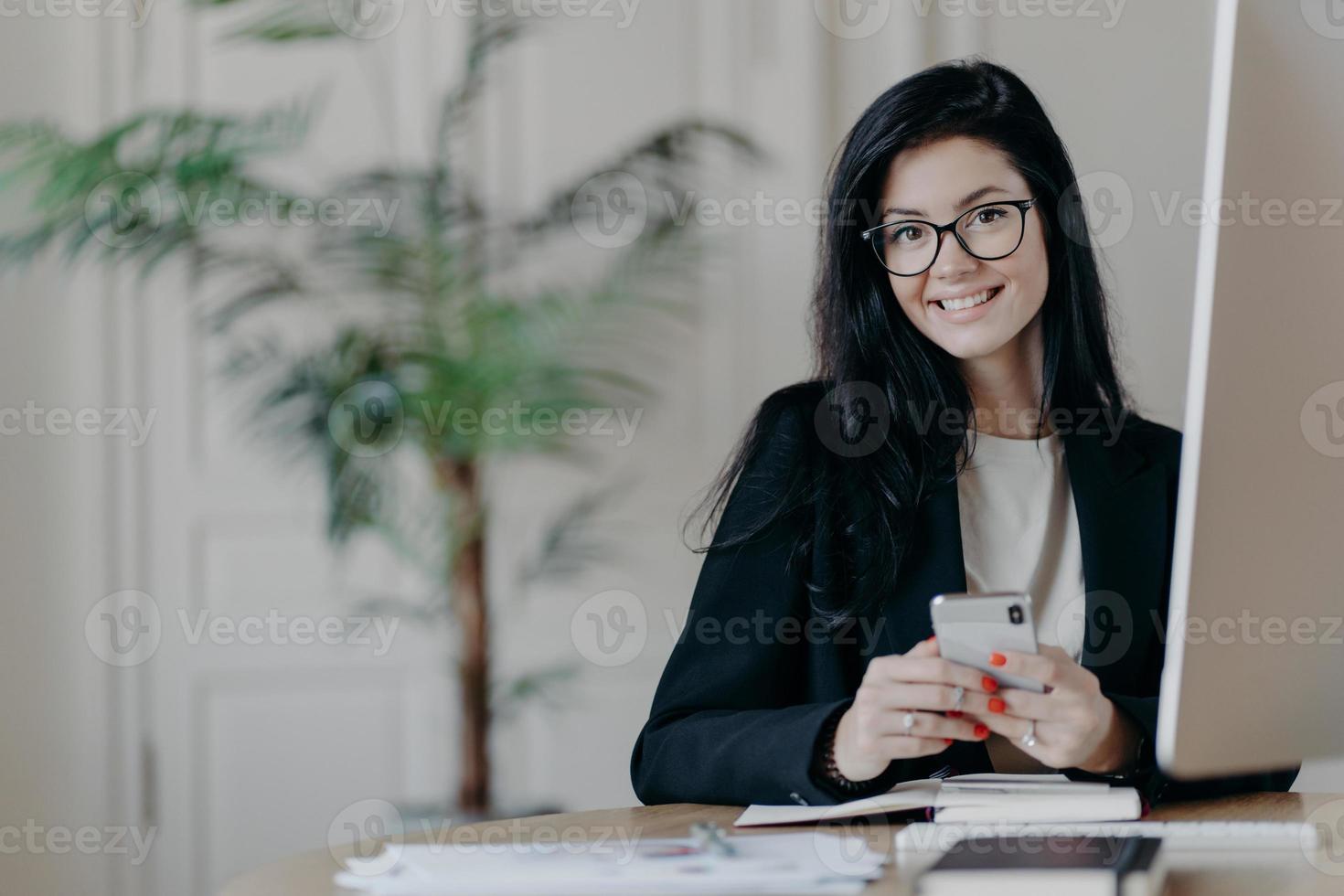 Happy female office worker in stylish black jacket browses smartphone with cheerful expression, sends text messages, works on important project, poses in coworking space. Modern technologies photo