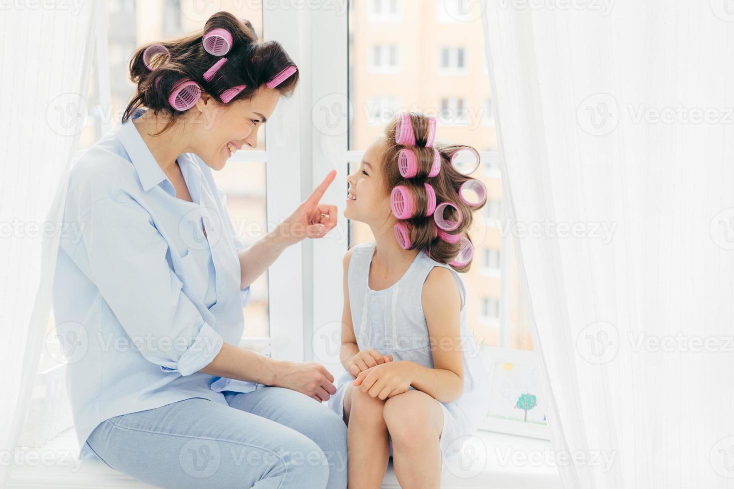 Cheerful two mother and daughter do hair nd have fun together, have curlers on head, sit on window sill. Affectionate mum touches nose of her small kid, prepare for party, care of hair and beauty photo