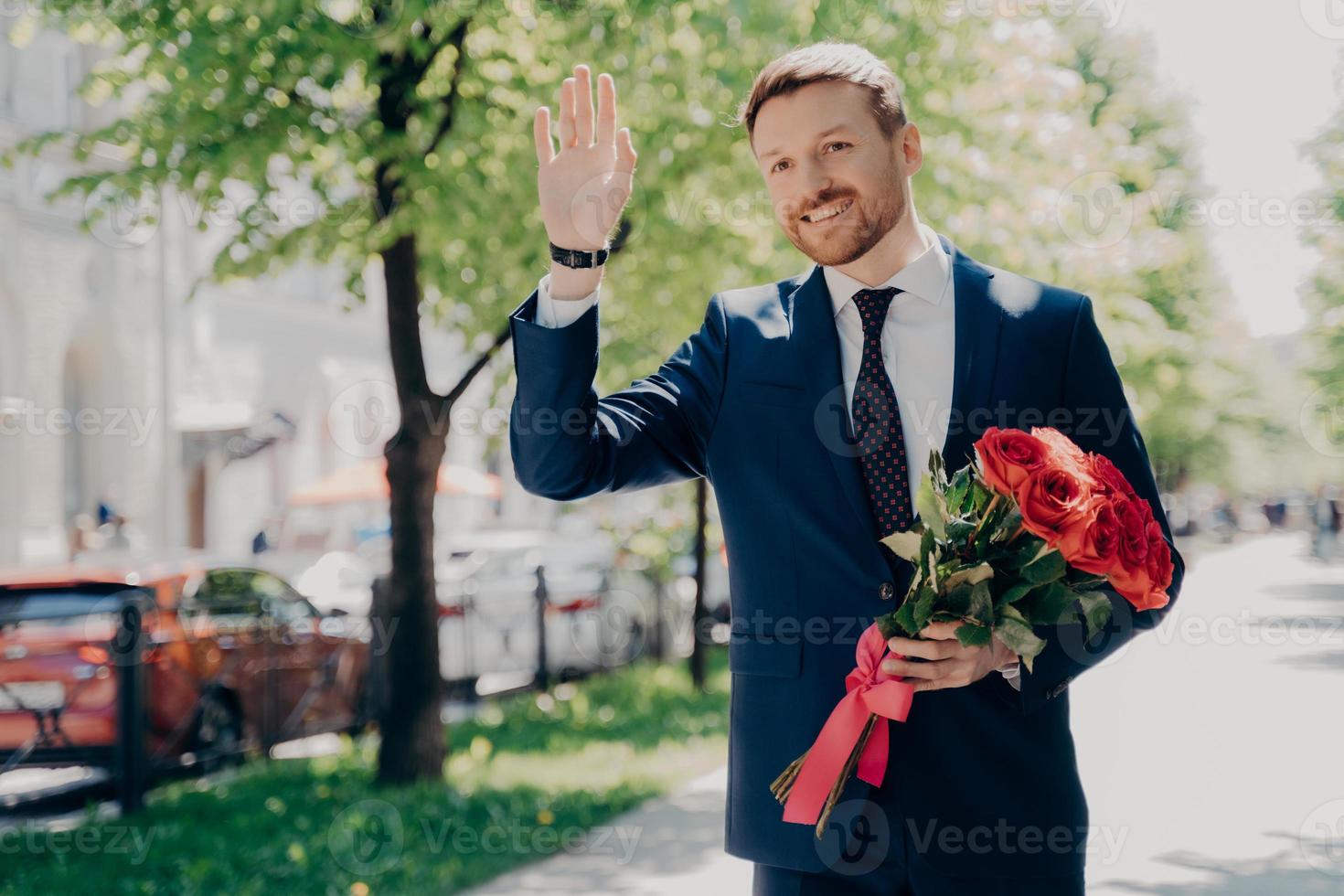 hombre de negocios feliz en traje con ramo saludando a alguien en el parque de la ciudad foto