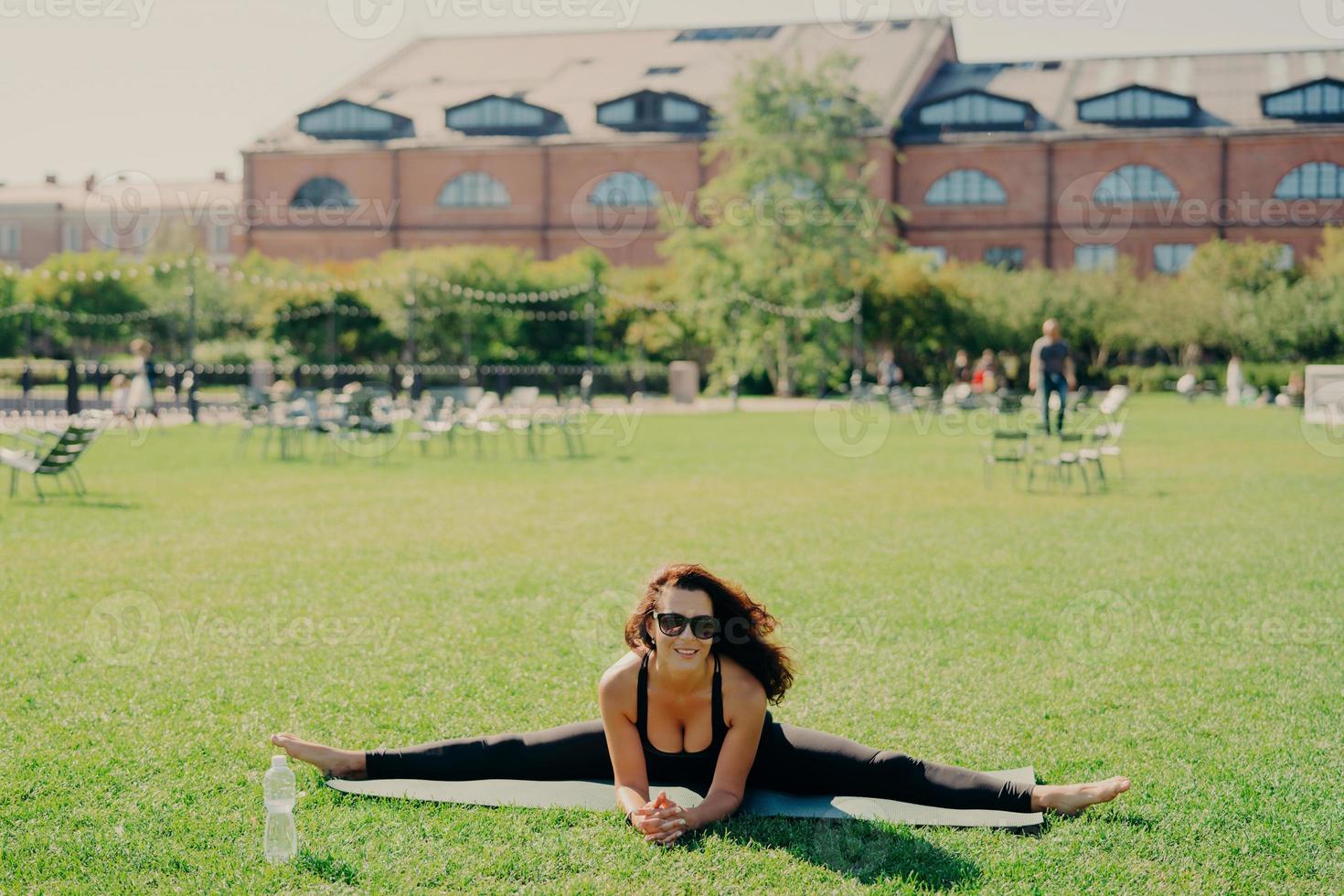 Fit active European woman does pilates on fitness mat wears sunglasses and sportswear shows great progress poses on green lawn has happy expression trains outside. Fitness and stretching concept photo