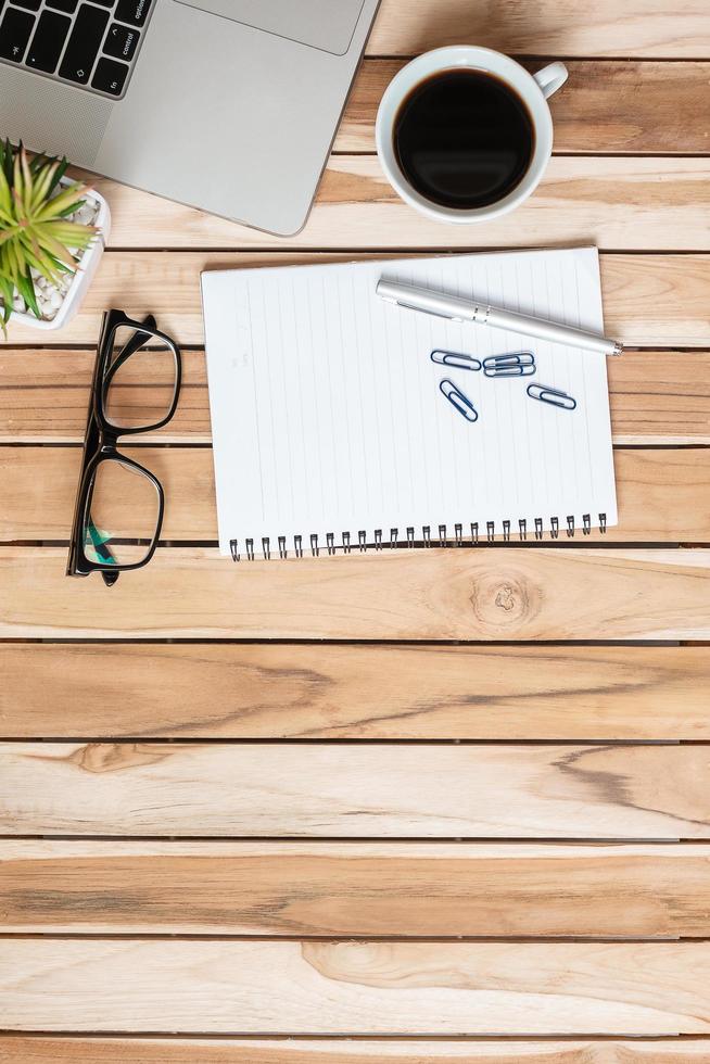 Top view Office desk with blank notebook, pen, paper clips, computer laptop, plant pot, eyeglasses and coffee cup on wood table background. workspace or home office with copy space for text concept photo