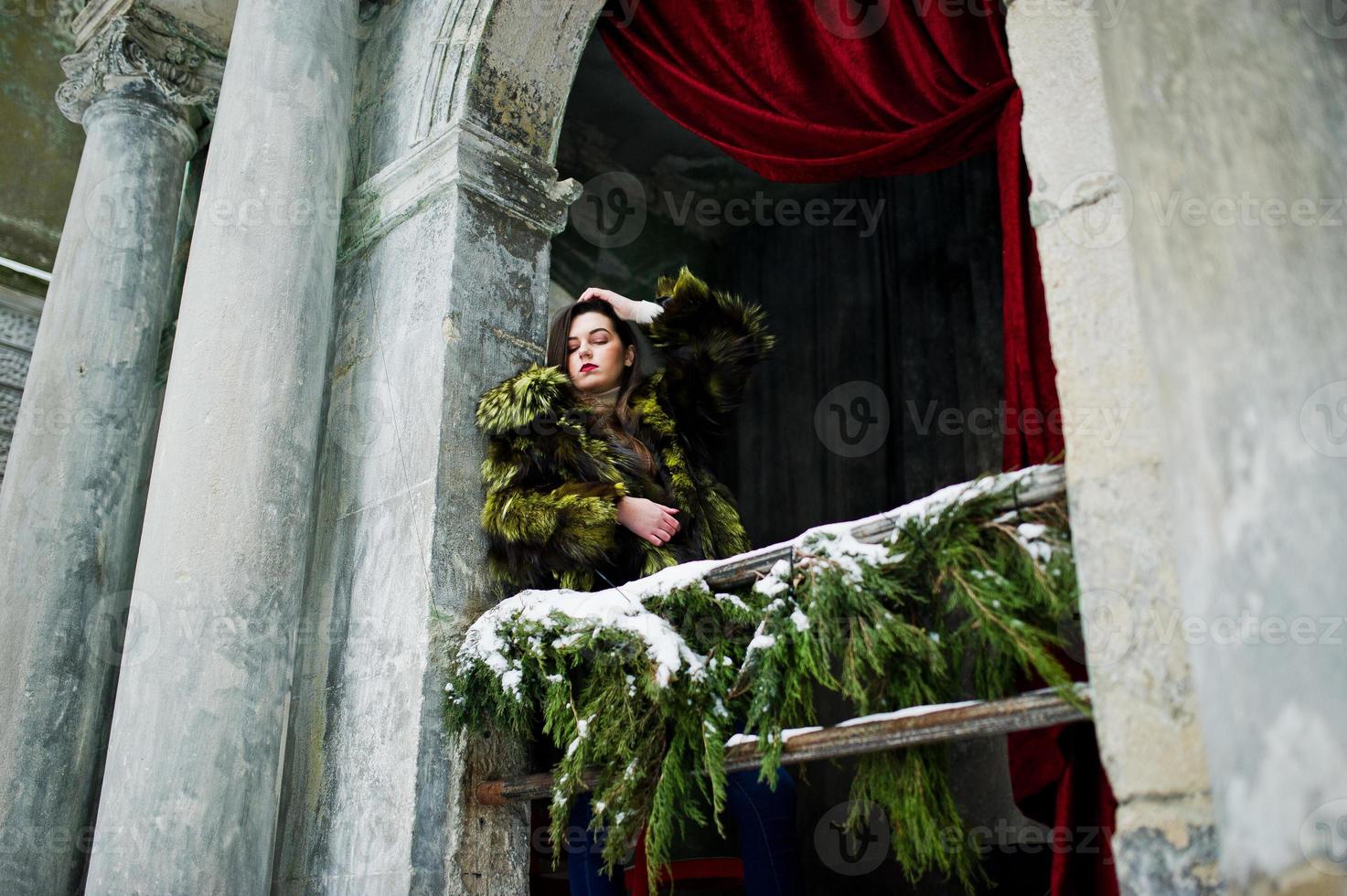 Brunette girl in green fur coat against old arch with columns and red curtains. photo