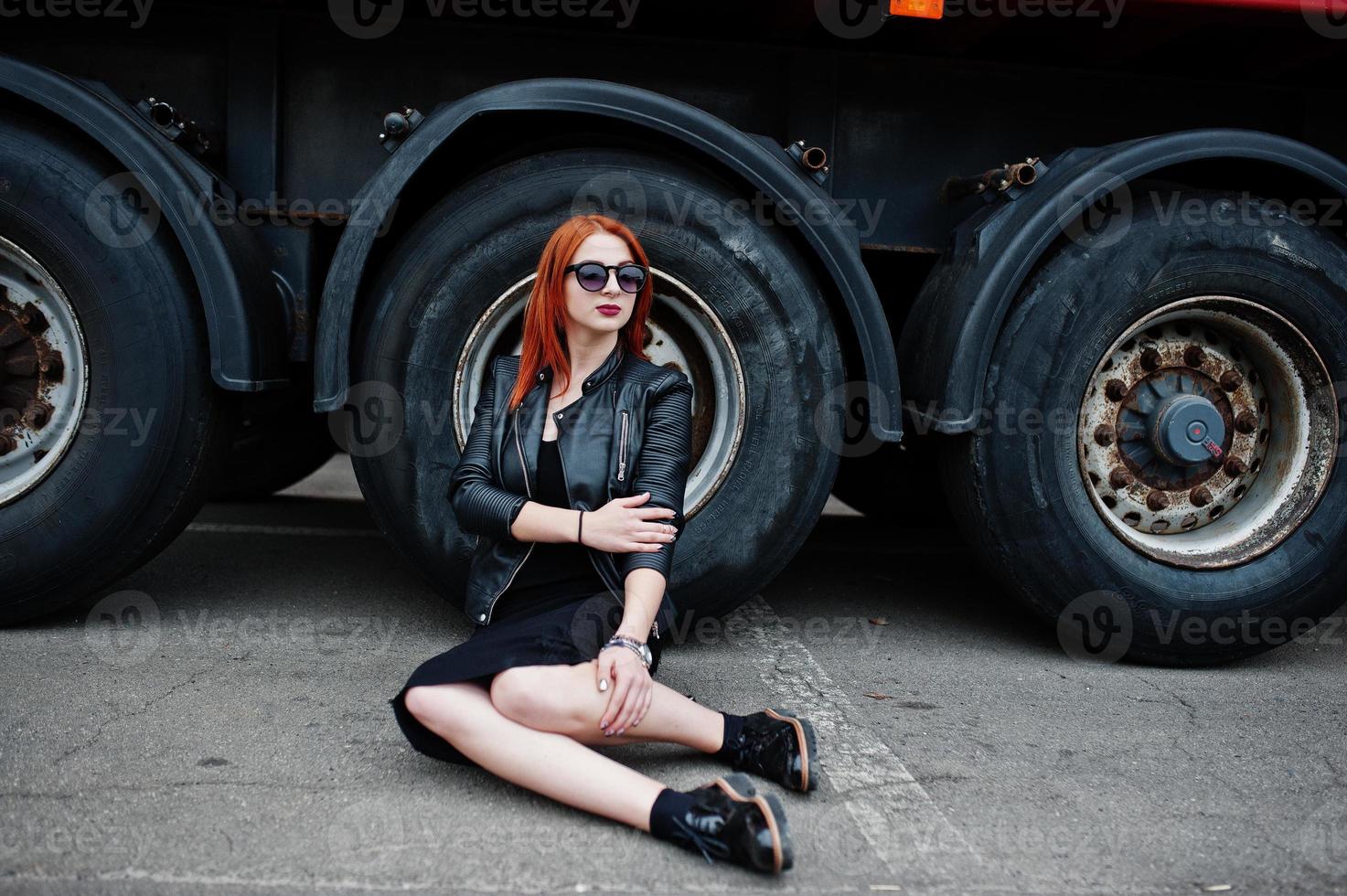 Red haired stylish girl wear in black, sitting against large truck wheels. photo