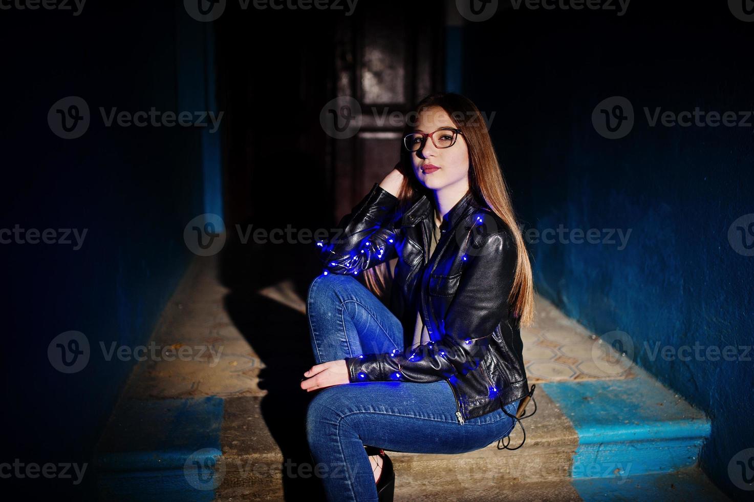 Night portrait of girl model wear on glasses, jeans and leather jacket, with blue garland on her. photo