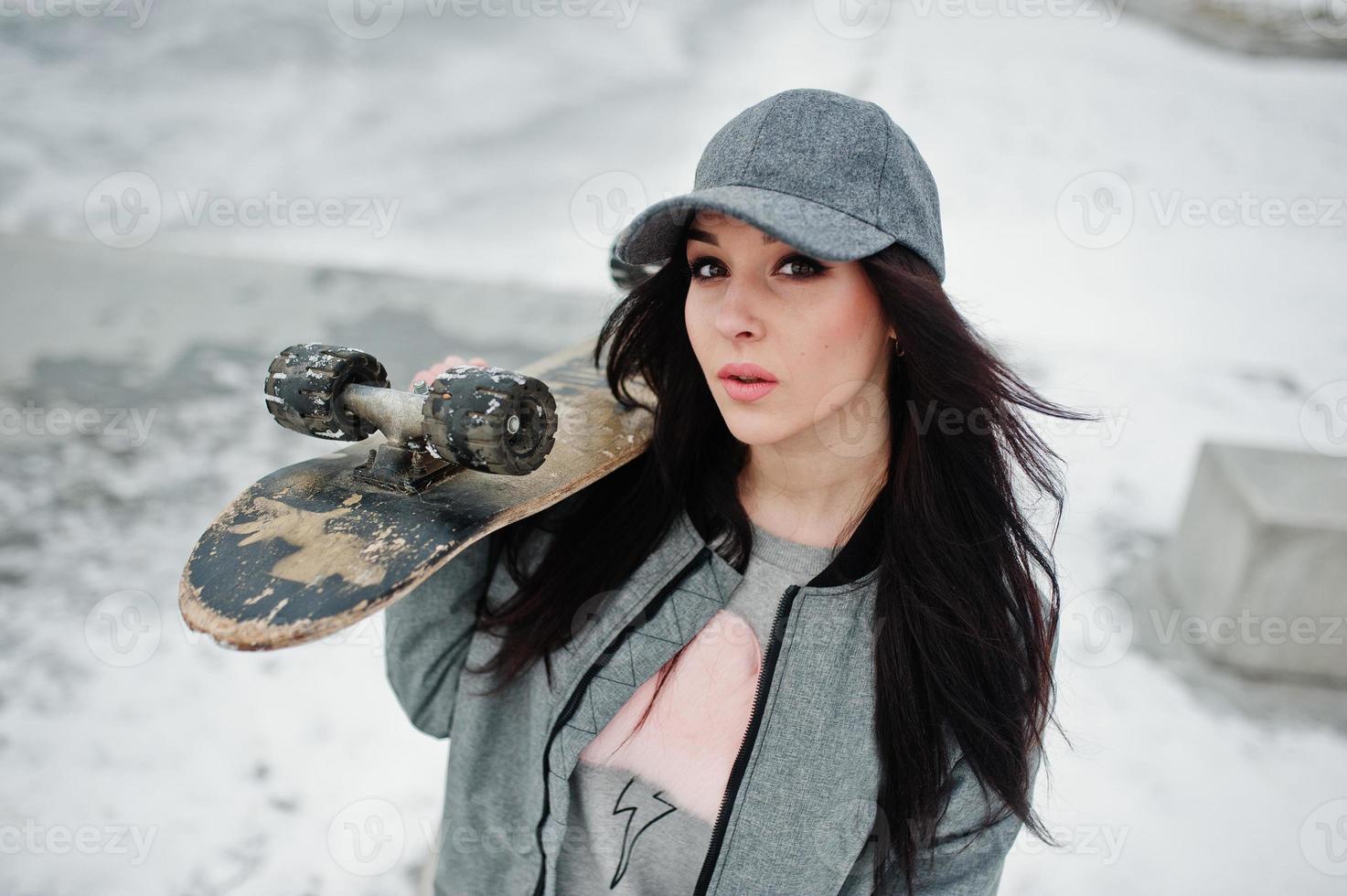 Stylish brunette girl in gray cap, casual street style with skate board on winter day. photo