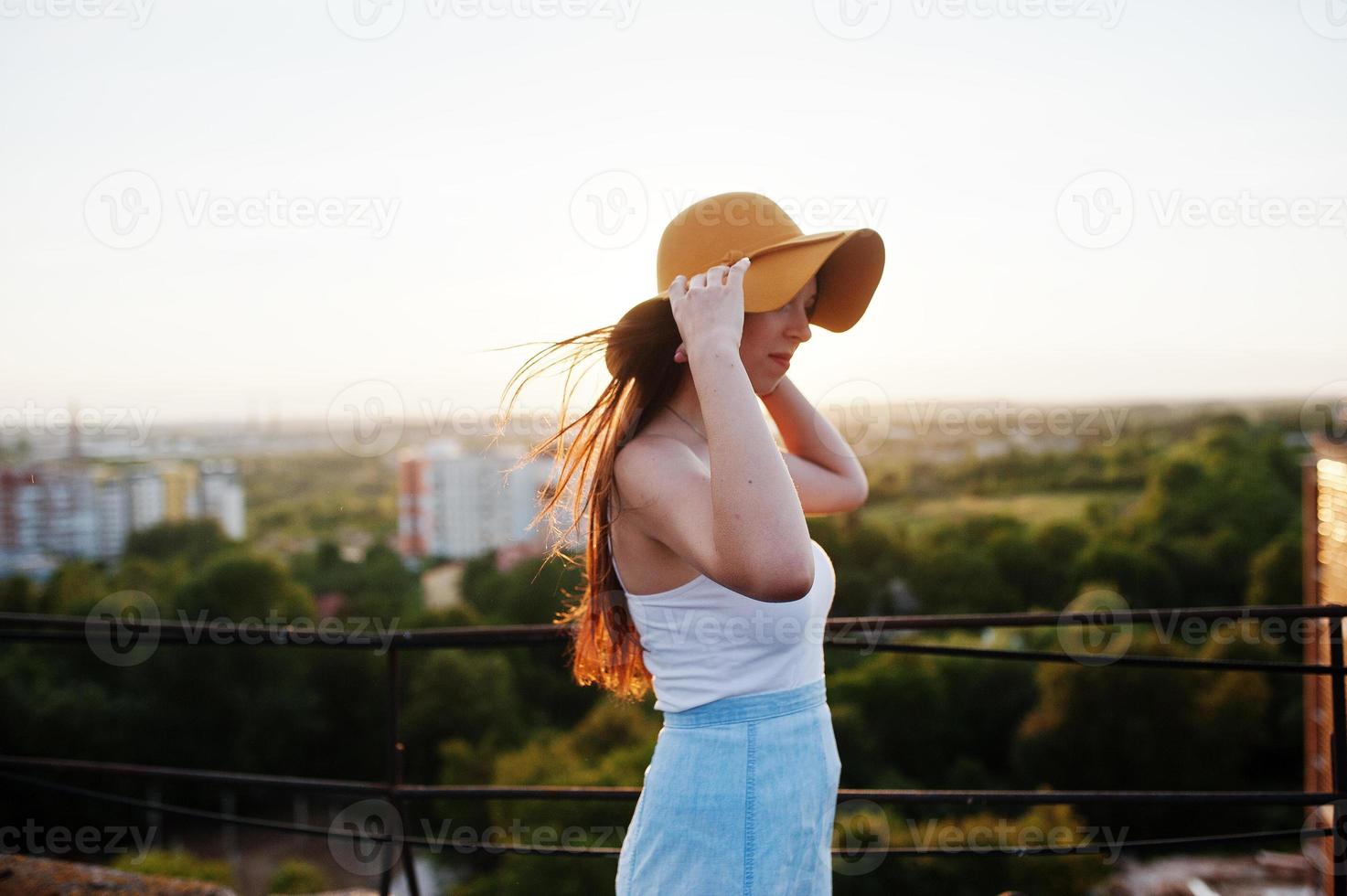 Portrait of a pretty young woman in white t-shirt and blue skirt posing on the rooftop with her orange hat at the sunset. photo