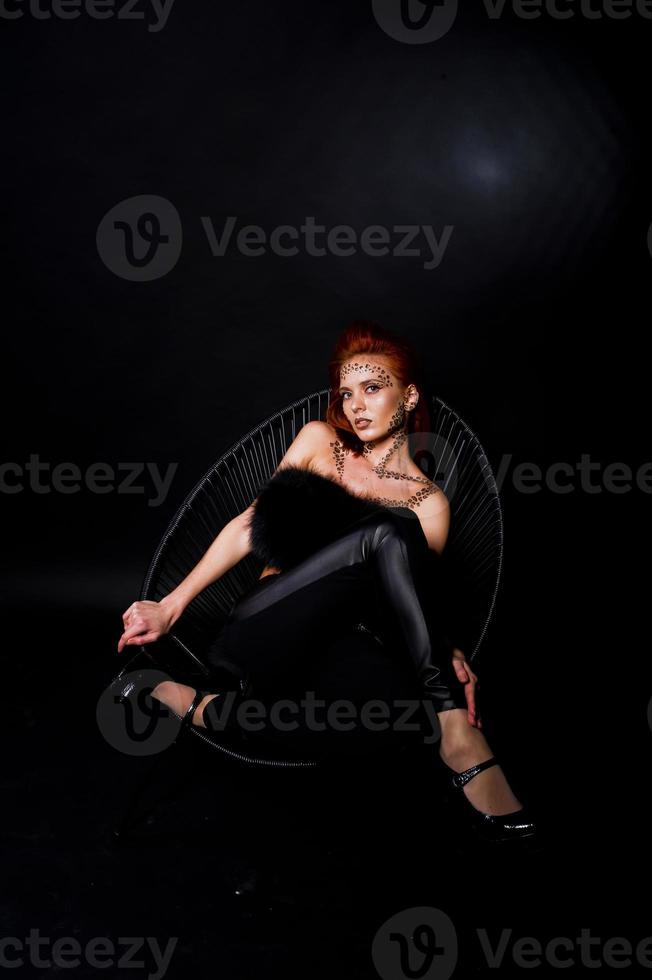 Fashion model red haired girl with originally make up like leopard predator isolated on black. Studio portrait on chair. photo