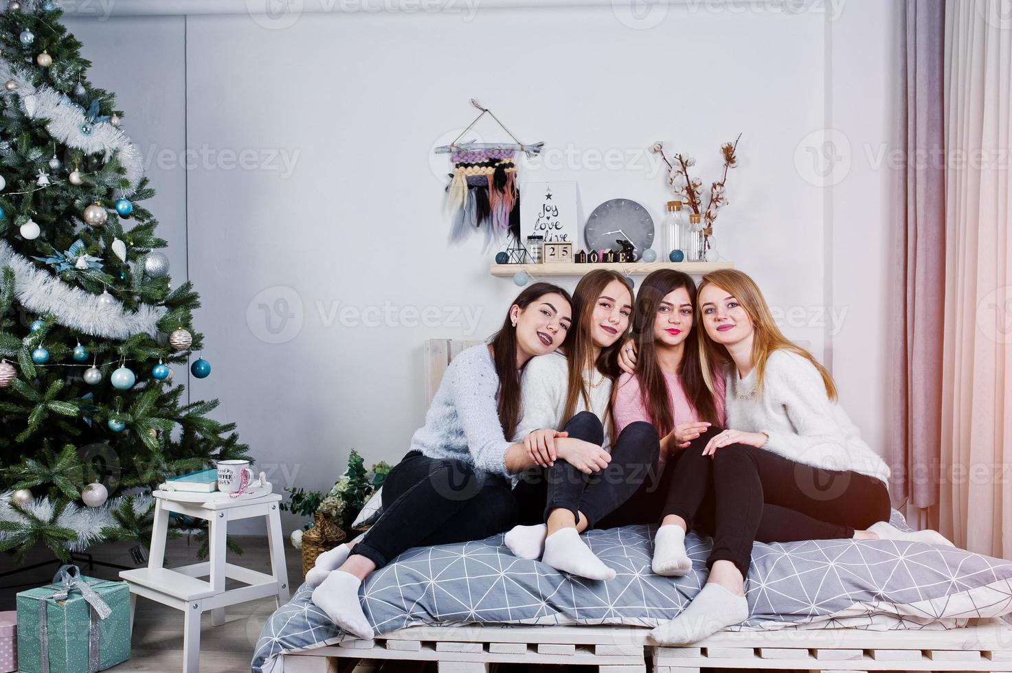 Four cute friends girls wear on warm sweaters and black pants on bed at new year decorated room on studio. photo
