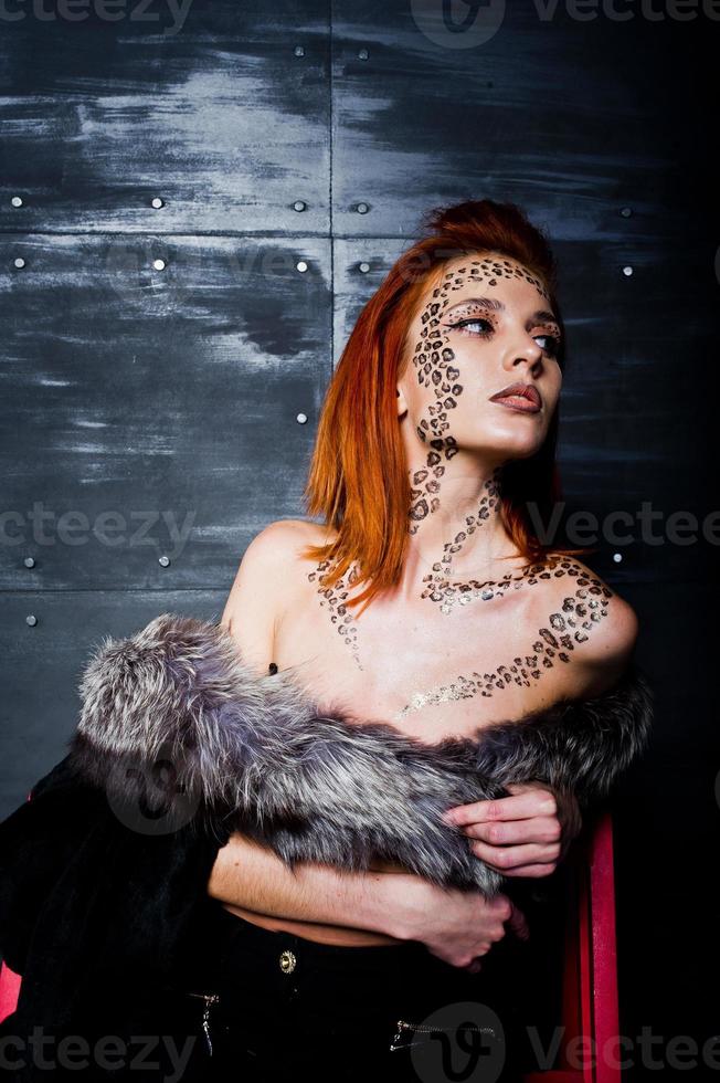 Fashion model red haired girl with originally make up like leopard predator against steel wall. Studio portrait on ladder. photo