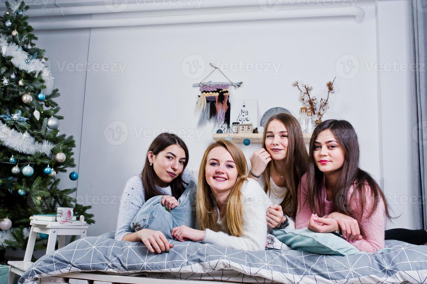 Four cute friends girls wear on warm sweaters and black pants on bed at new year decorated room on studio, play with pillows. photo