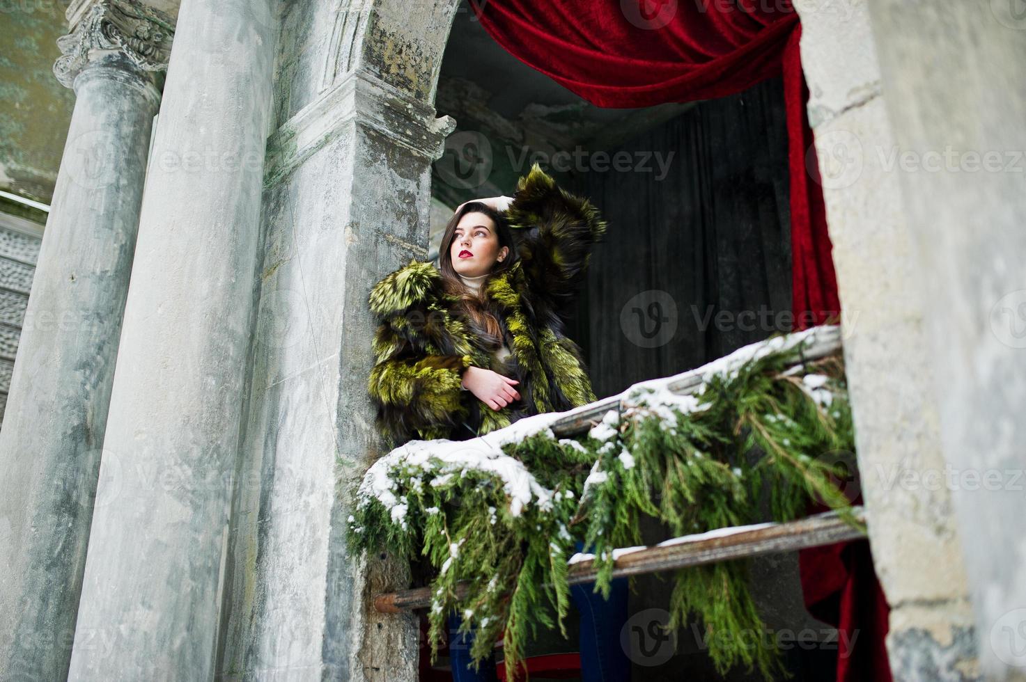 Brunette girl in green fur coat against old arch with columns and red curtains. photo