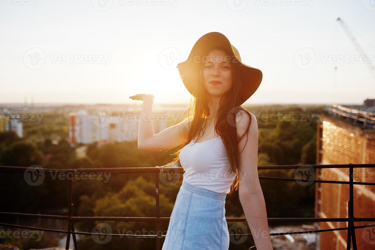 Portrait of a flawless young woman in white t-shirt, blue skirt and orange hat pointing at the sun with her hand while standing on the rooftop. photo