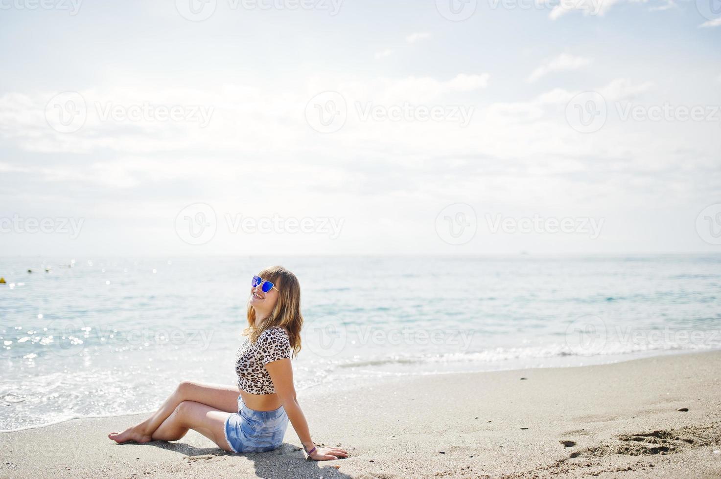 Beautiful model relaxing on a beach of sea, wearing on jeans short, leopard shirt and sunglasses. photo