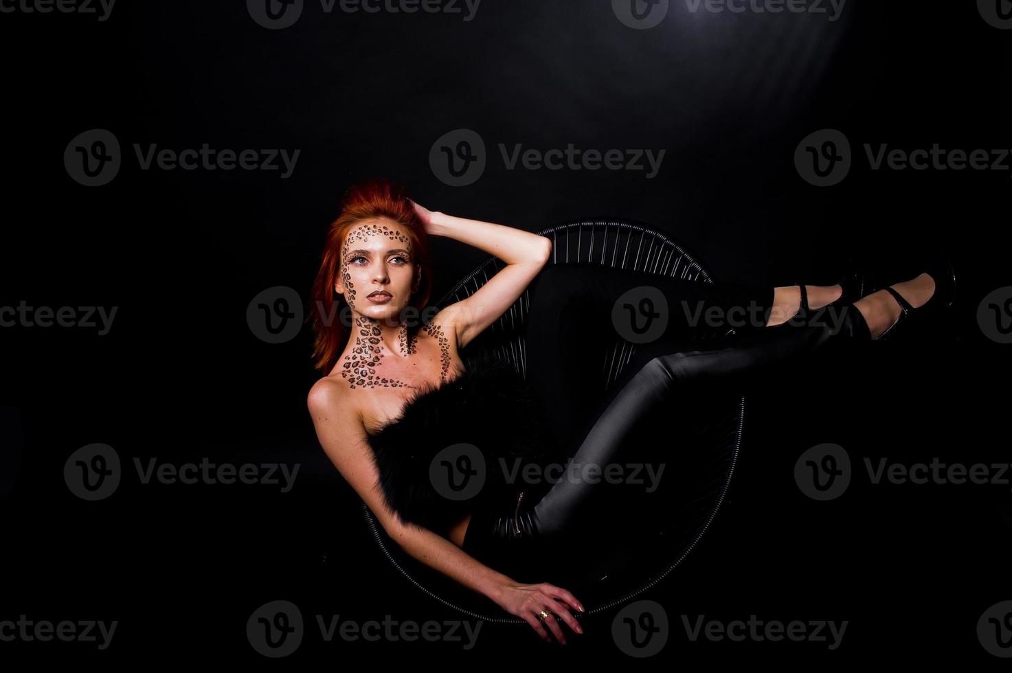 Fashion model red haired girl with originally make up like leopard predator isolated on black. Studio portrait on chair. photo