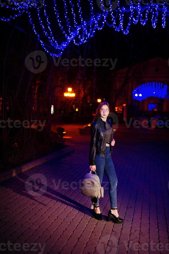 retrato nocturno de modelo de niña con gafas, jeans y chaqueta de cuero, con mochila en las manos, contra la guirnalda de luces azules de la calle de la ciudad. foto