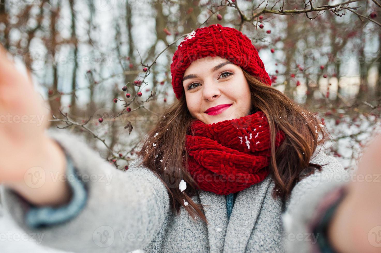 Portrait of gentle girl in gray coat , red hat and scarf near the branches of a snow-covered tree, holding camera for selfie. photo