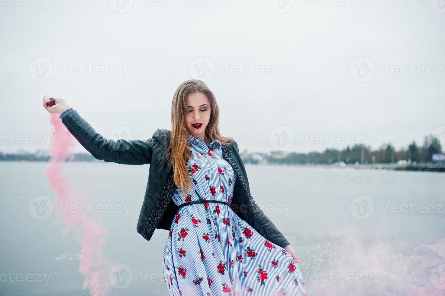 chica elegante con chaqueta de cuero sostiene una bengala de humo rosa en el día de invierno contra el lago congelado. foto