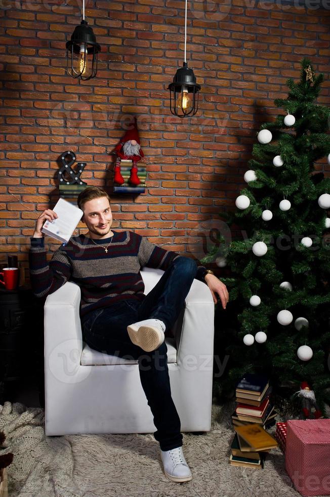 retrato de estudio de un hombre con un libro sentado en una silla contra un árbol de navidad con adornos. foto