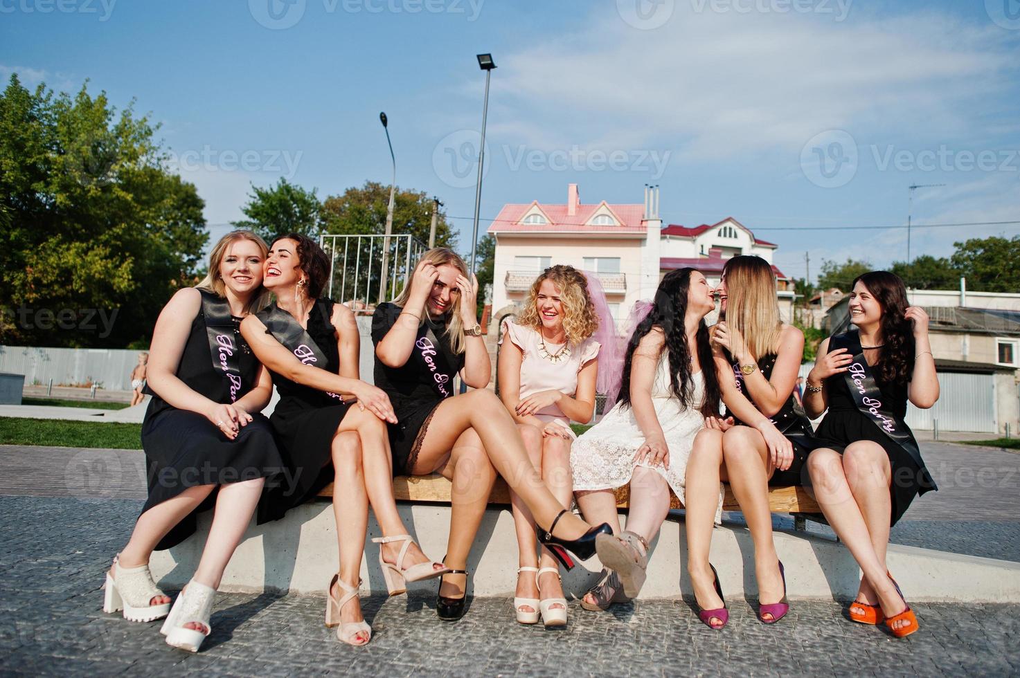 Group of 7 girls wear on black and 2 brides sitting on bench and having fun at hen party. photo