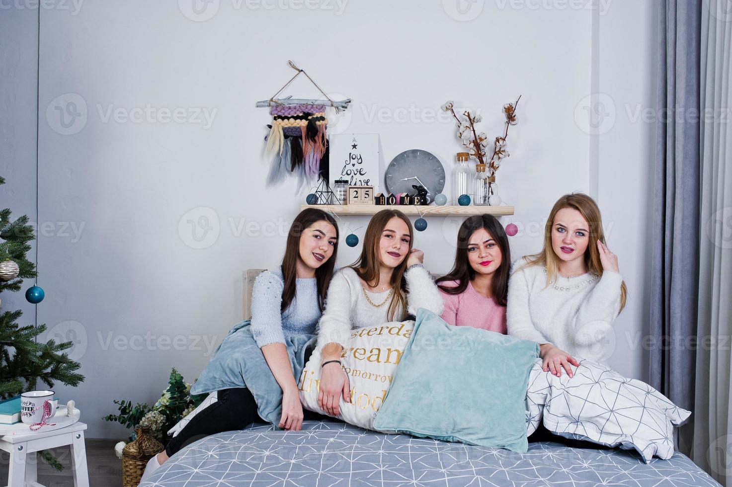 Four cute friends girls wear on warm sweaters and black pants on bed at new year decorated room on studio, play with pillows. photo