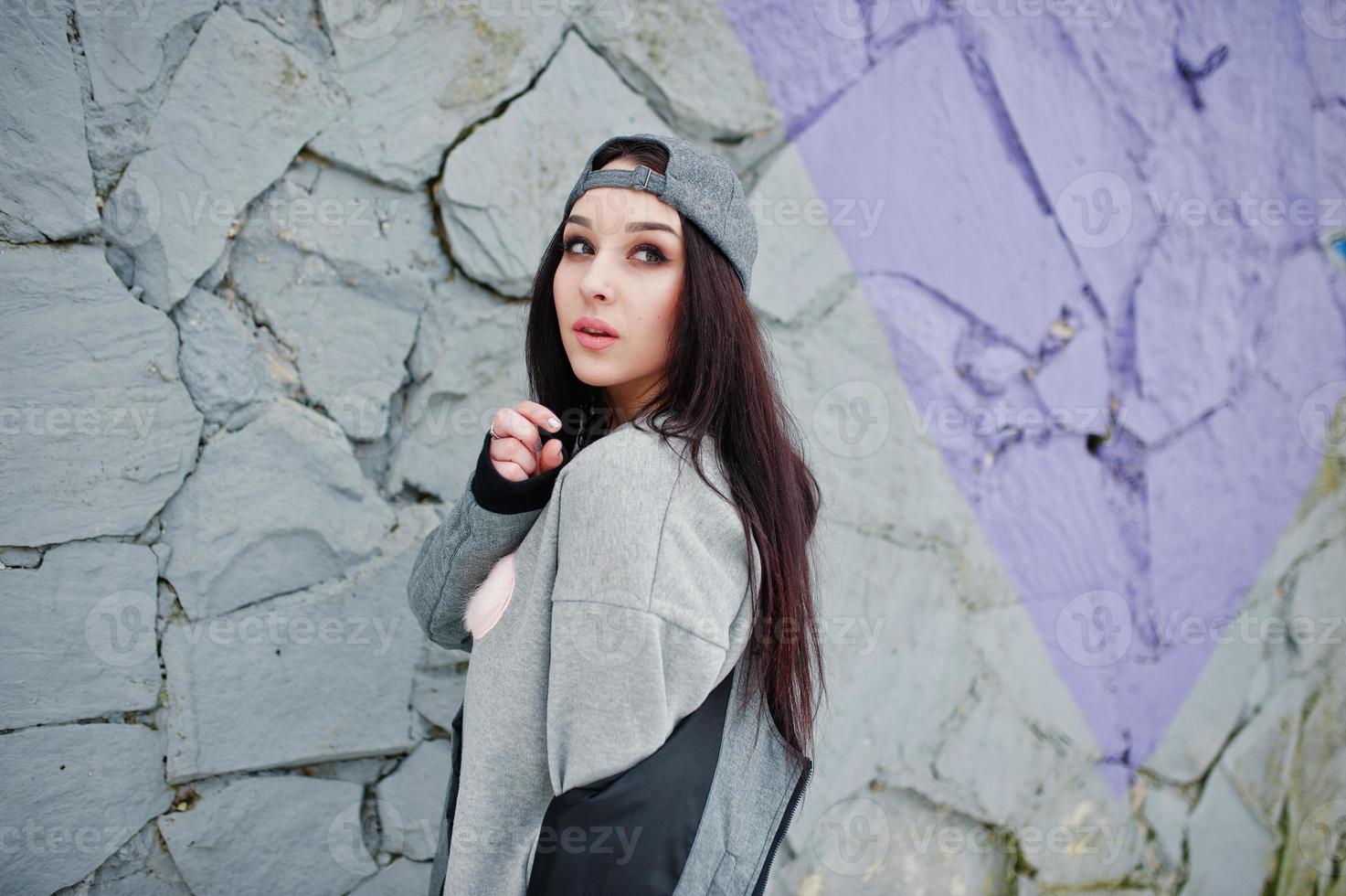 Stylish brunette girl in gray cap, casual street style on winter day against colored wall. photo