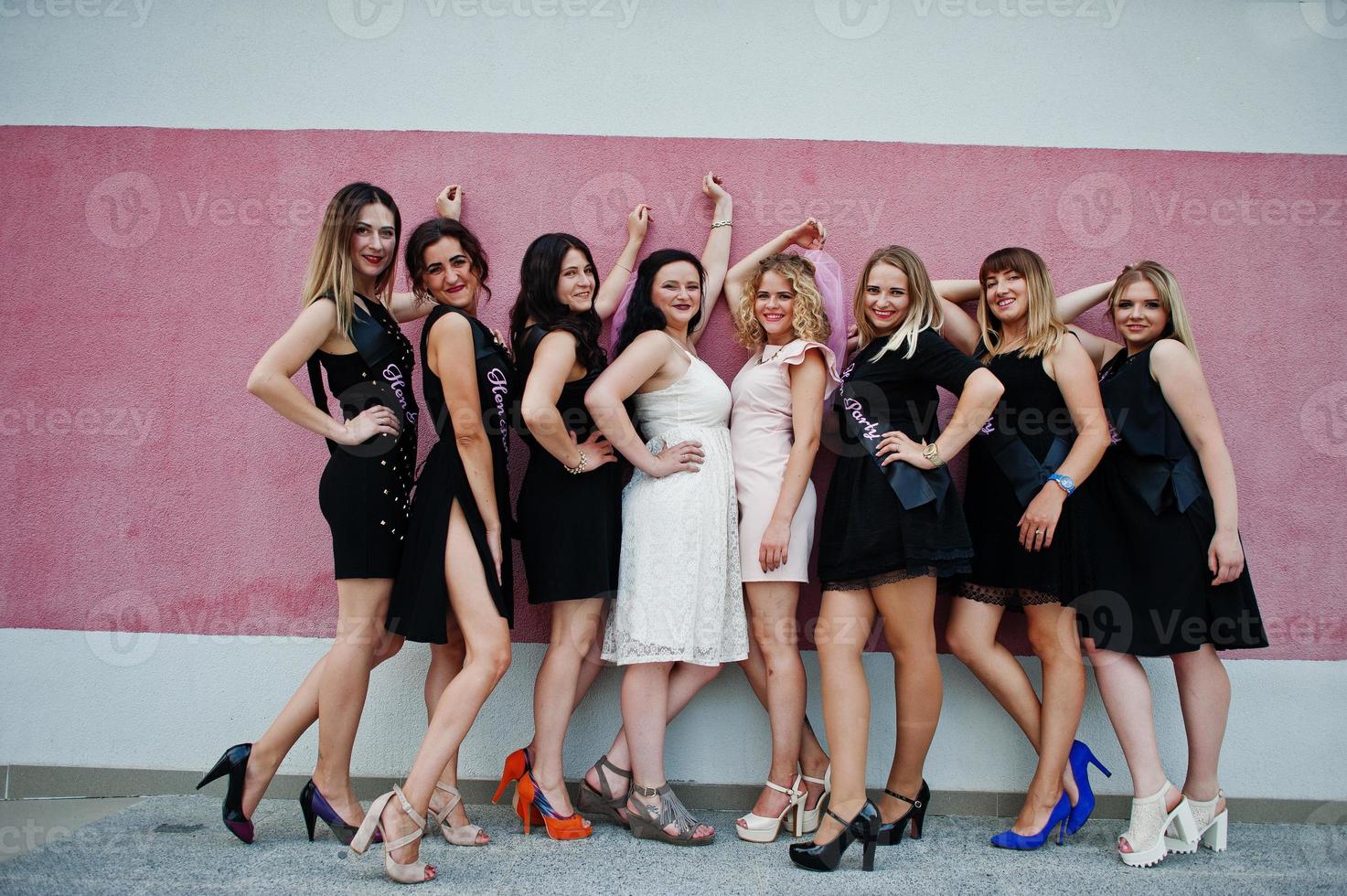 Group of 8 girls wear on black and 2 brides at hen party posed against pink wall. photo