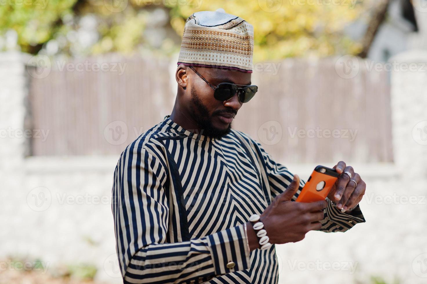 Portrait of stylish black african american man at hat and sunglasses against sunny autumn fall background doing selfie from mobile phone. Rich people in africa at traditional dress. photo