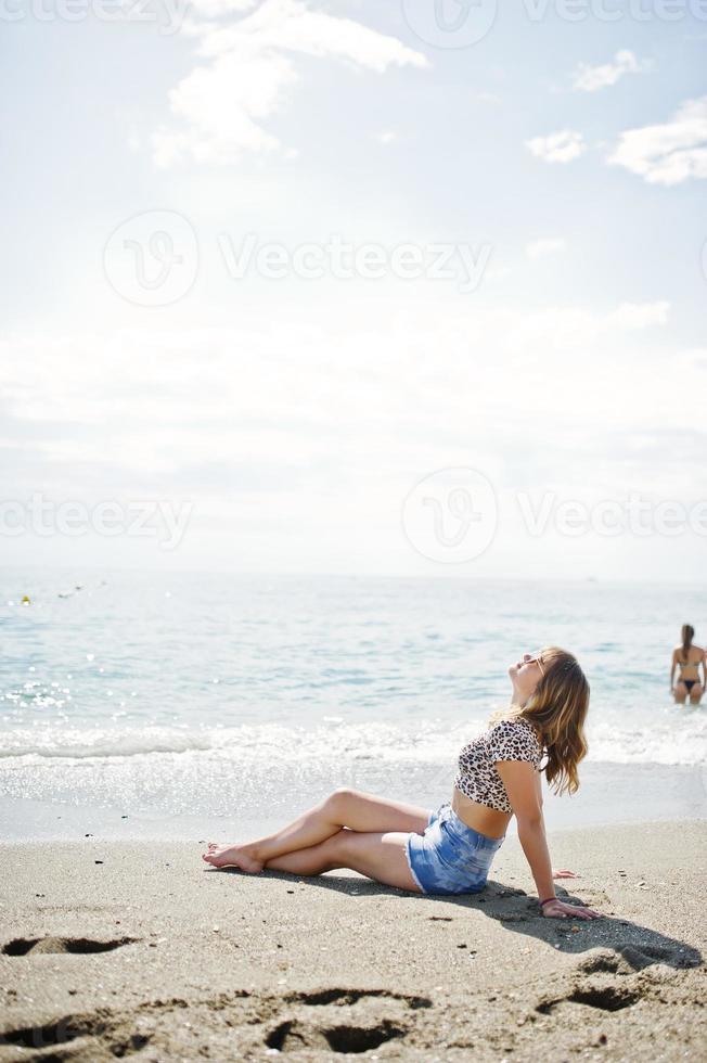 hermosa modelo relajándose en una playa de mar, usando jeans cortos, camisa de leopardo y gafas de sol. foto