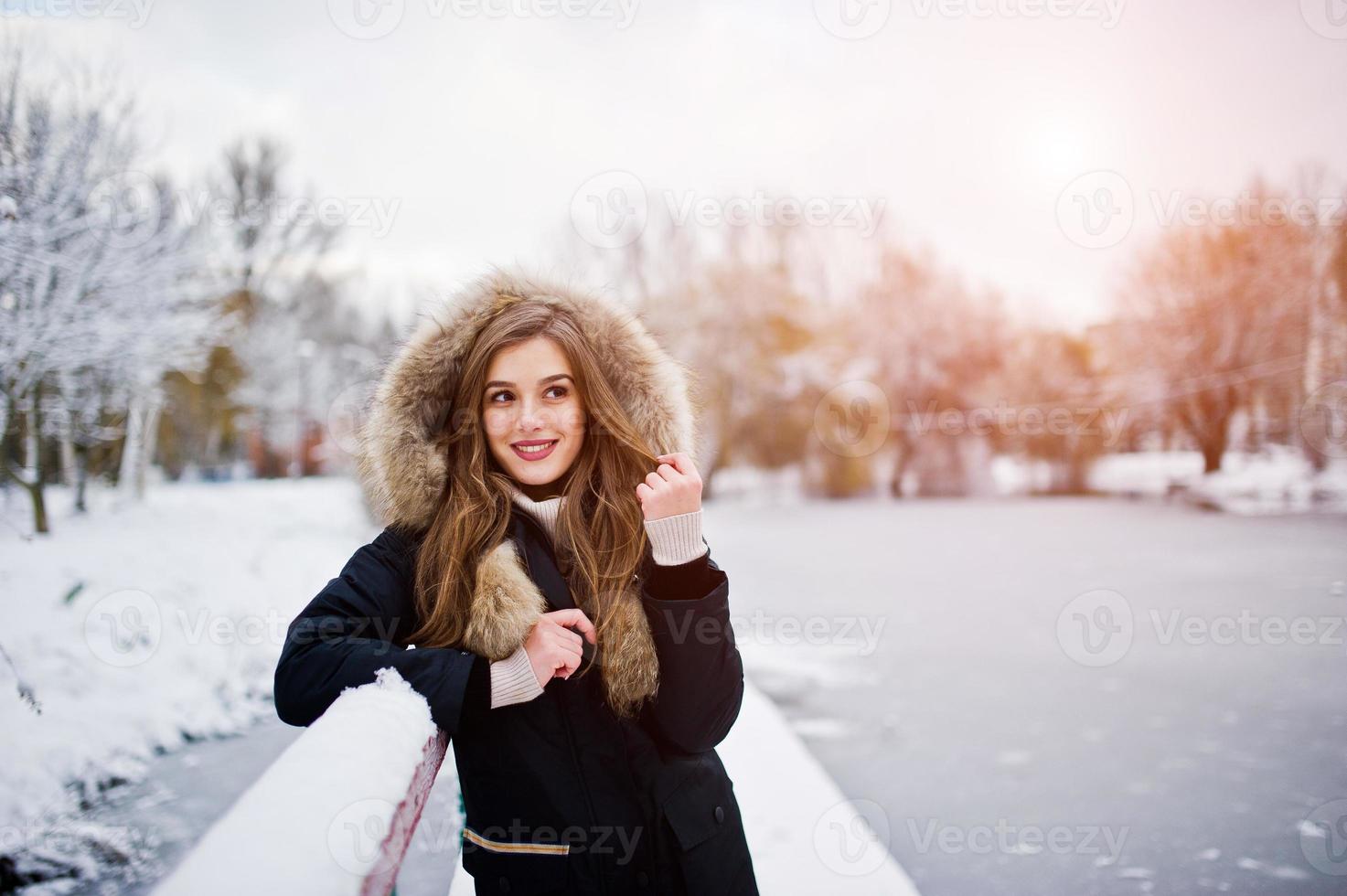 Beautiful brunette girl in winter warm clothing. Model on winter jacket against frozen lake at park. photo