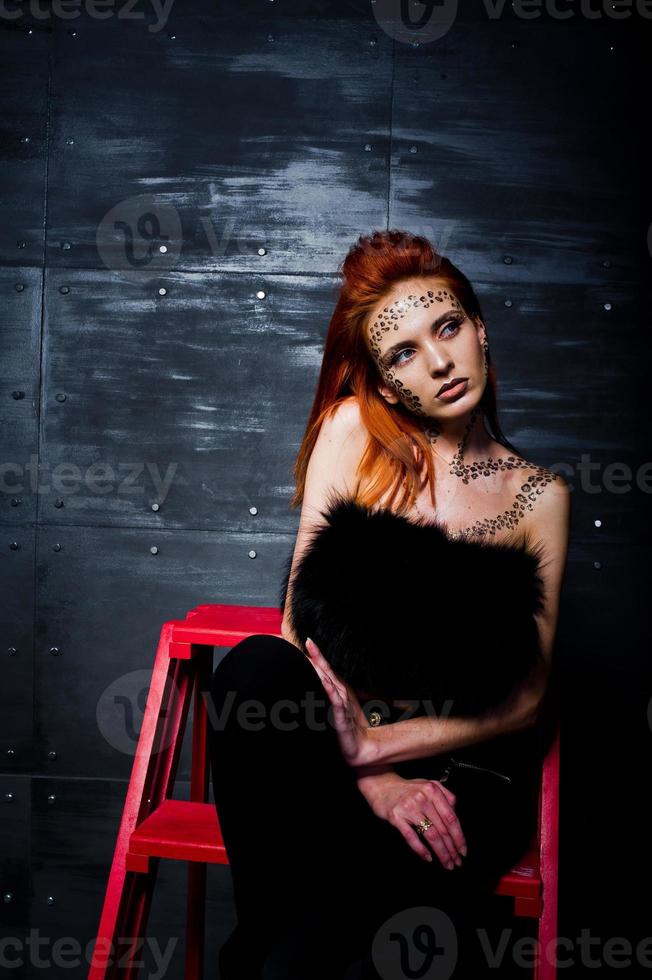 Fashion model red haired girl with originally make up like leopard predator against steel wall. Studio portrait on ladder. photo