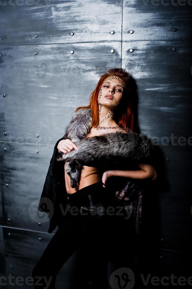 Fashion model red haired girl with originally make up like leopard predator wear on furs against steel wall. Studio portrait. photo