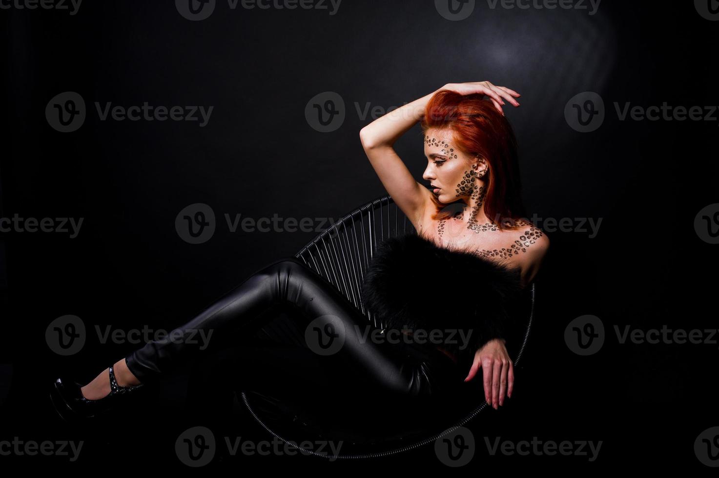 Fashion model red haired girl with originally make up like leopard predator isolated on black. Studio portrait on chair. photo