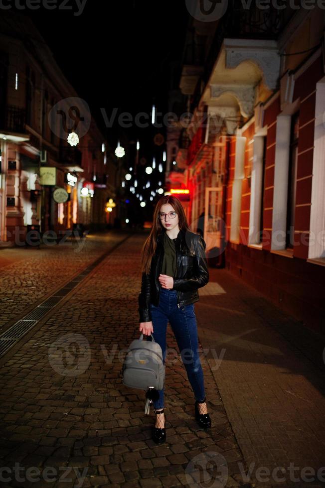 retrato nocturno de modelo de niña con gafas, jeans y chaqueta de cuero, con mochila, contra las luces de las calles de la ciudad. foto